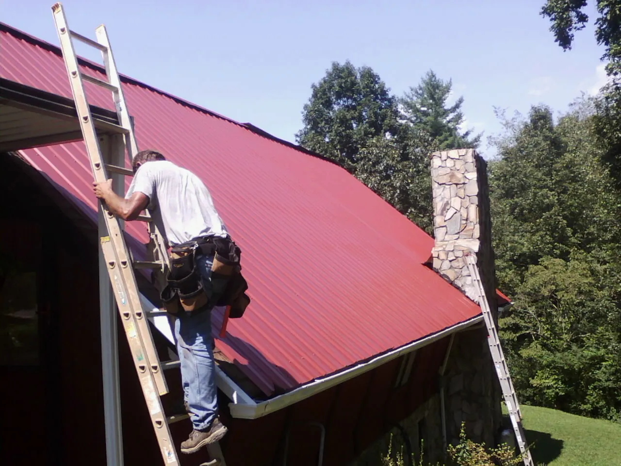 Roofer on a ladder beside a red metal roof and stone chimney — Soffit & Fascia Repair by Roofing Brothers in Pauma Valley